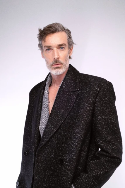 Headshot of a mature adult man with grey hair and a beard, wearing a dark textured coat over a grey textured jacket. Light background.