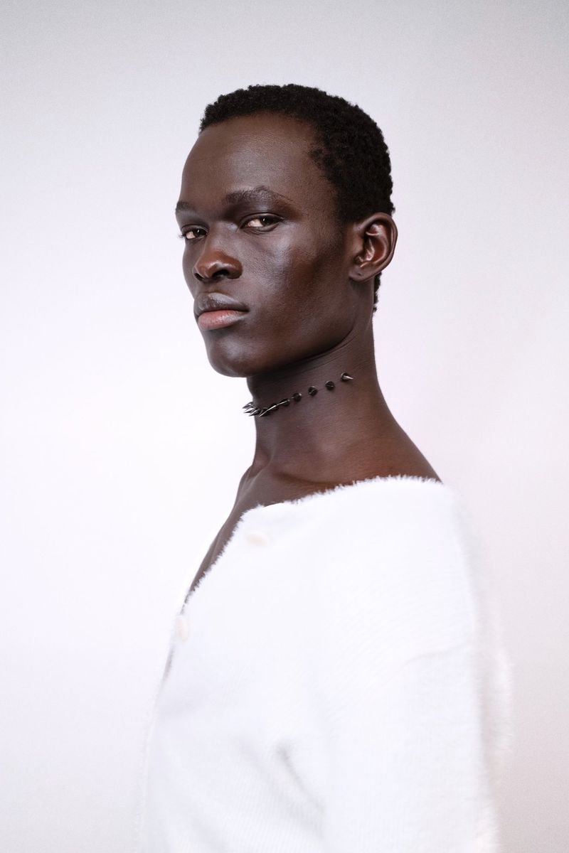 Portrait of a black male model in a white textured top wearing a dark spike necklace against a white backdrop.