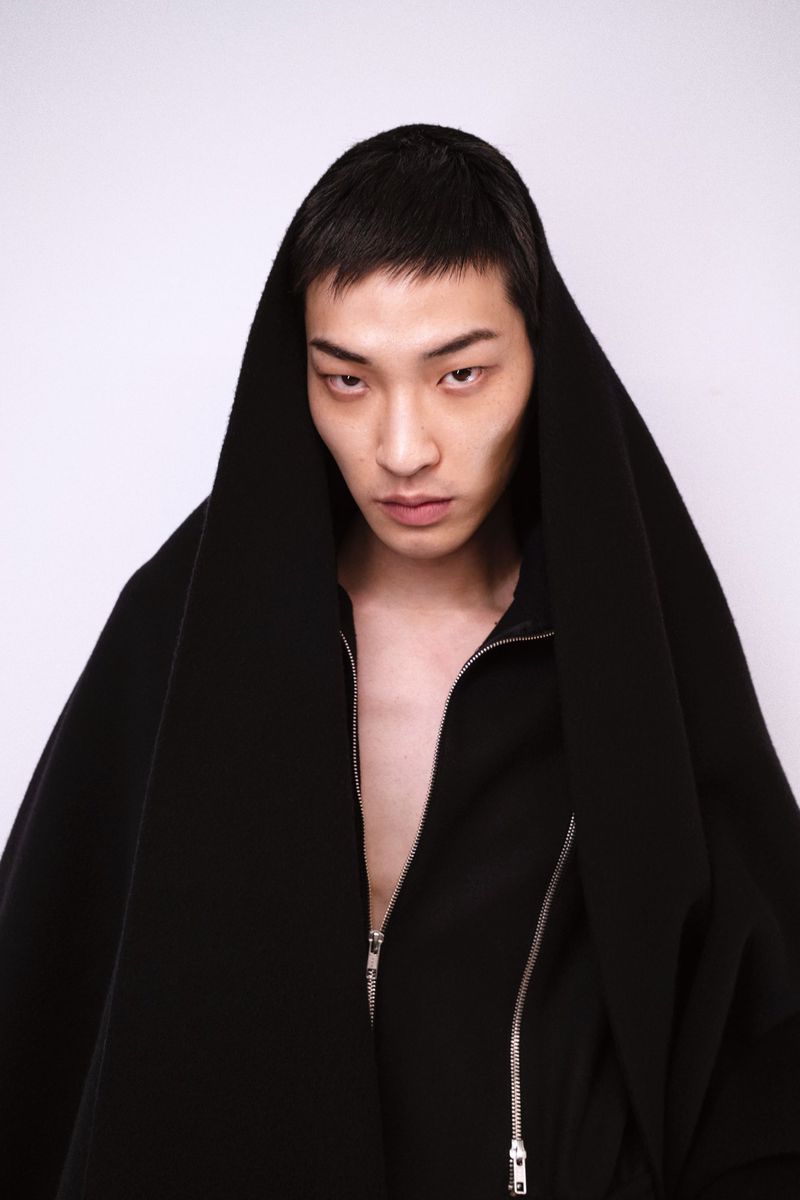 Asian male model wearing a large black hooded zippered jacket poses against a white background in a portrait studio shot.