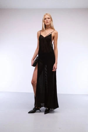 Model wearing a sheer black slip dress and pointed shoes standing against a grey seamless studio backdrop.