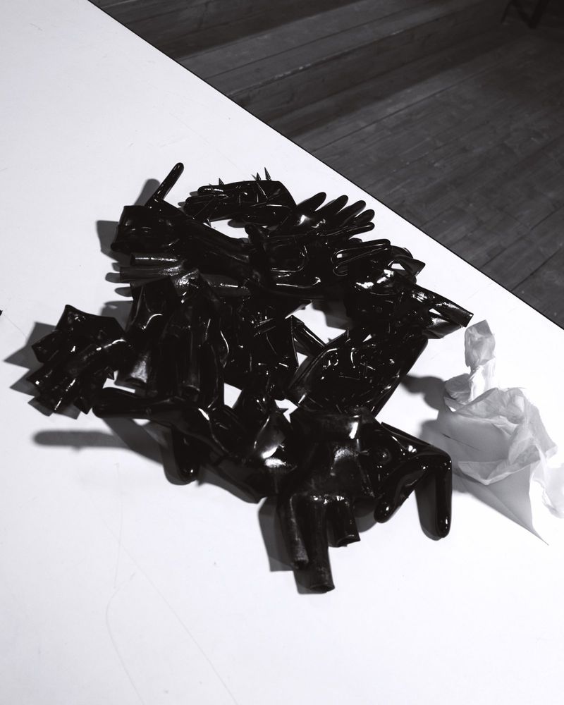 Monochrome close-up still life of shiny, dark gloves scattered on a white surface, contrasting with dark wooden floorboards in the background.