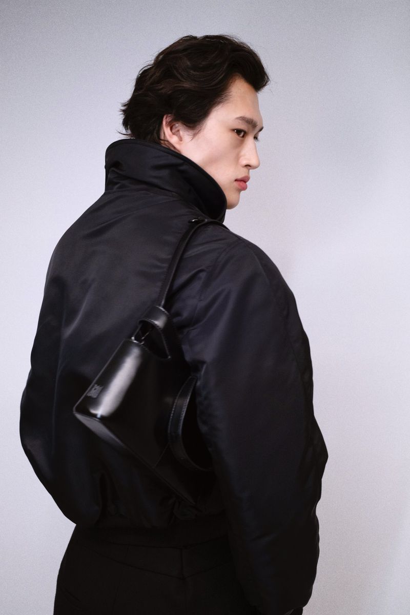 Model in a black jacket carrying a shoulder bag against a white seamless background