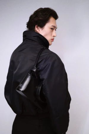 Model in a black jacket carrying a shoulder bag against a white seamless background