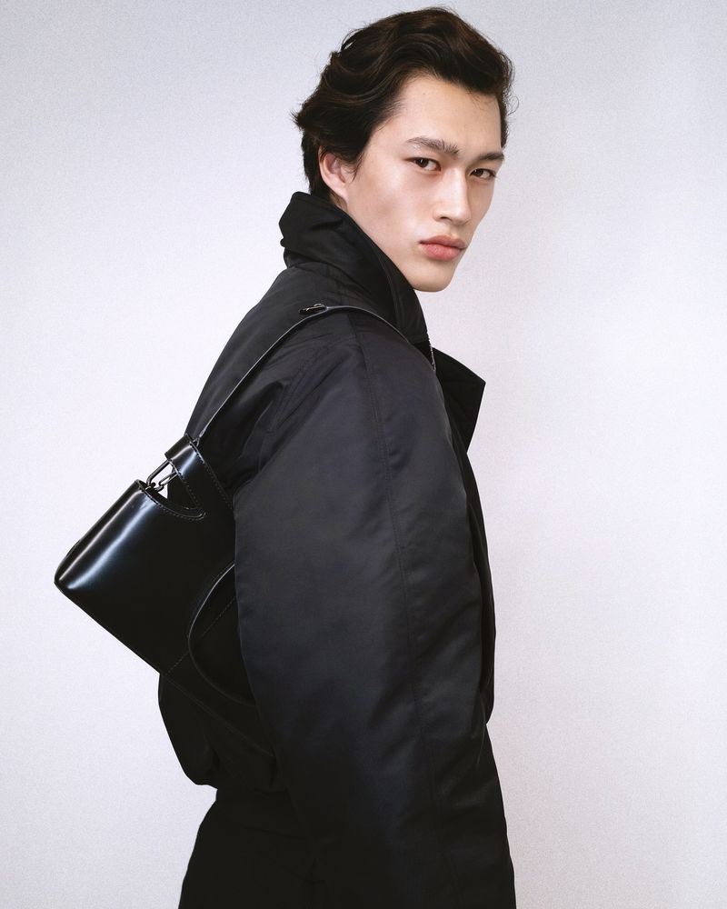 Portrait of a stylish young Asian man wearing a dark jacket and carrying a sleek black bag against a plain white backdrop in a studio.