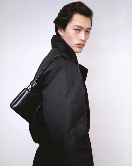 Portrait of a stylish young Asian man wearing a dark jacket and carrying a sleek black bag against a plain white backdrop in a studio.