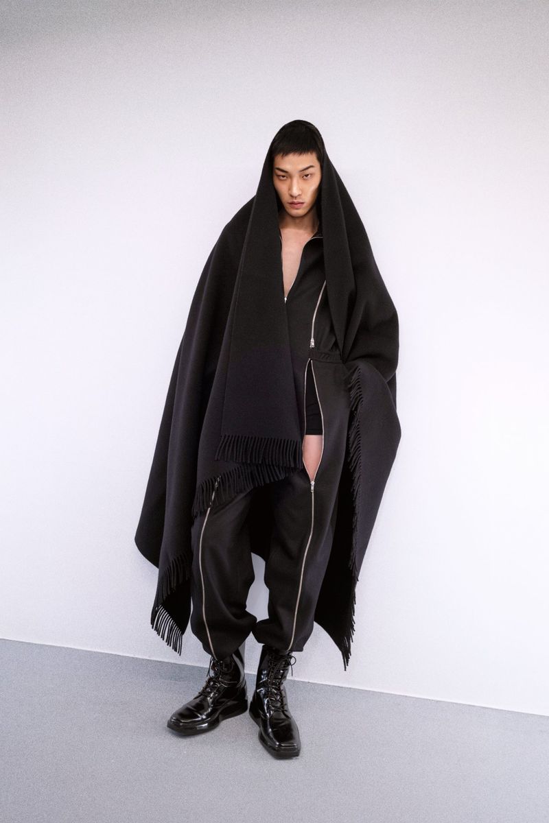 An Asian male model poses in a studio, wearing a black poncho with fringe, a black jumpsuit, and patent leather lace-up boots in a studio.
