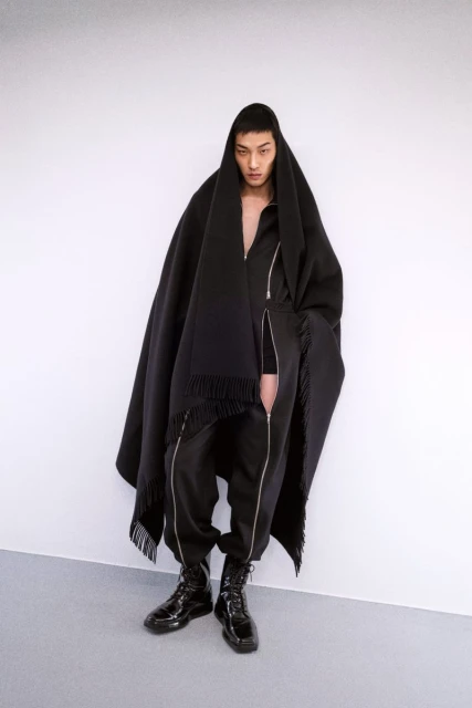 An Asian male model poses in a studio, wearing a black poncho with fringe, a black jumpsuit, and patent leather lace-up boots in a studio.