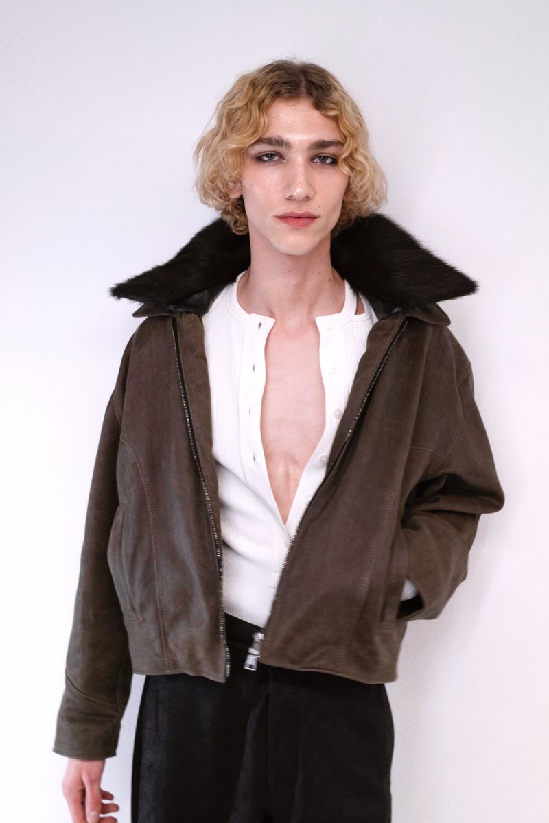 Model with blonde curly hair wearing a brown jacket with black fur collar and white henley shirt against a plain background