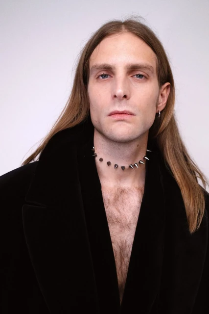 Portrait of an adult male with long hair wearing a dark coat and studded necklace against a white background in a photo studio.