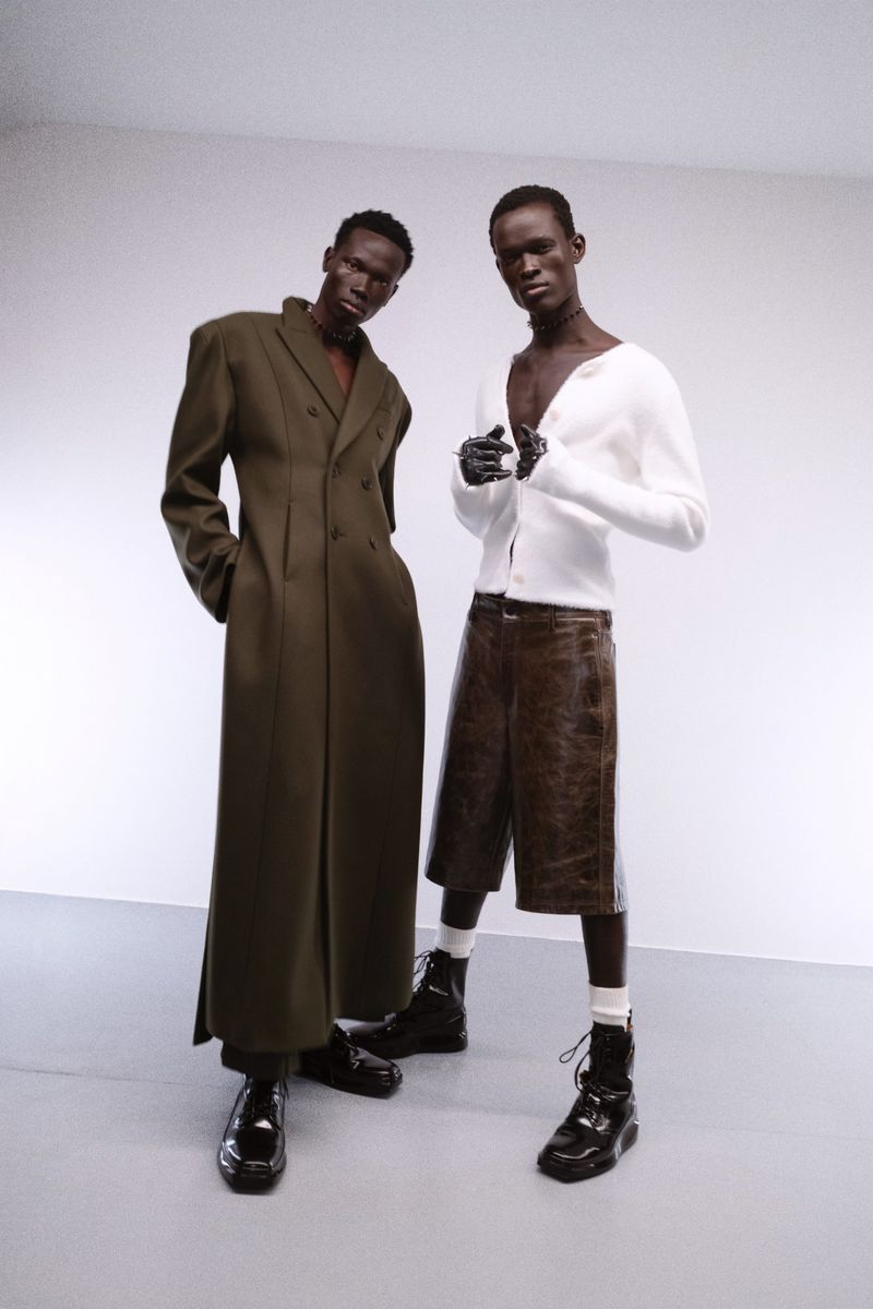 Two black male models pose in a minimalist studio, one in an olive trench coat and the other in a white cardigan and leather shorts.