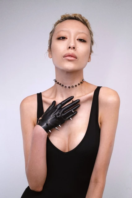 Woman in black dress and spiked glove posing in a studio with a light grey background, wearing a spiked necklace, eye-level, medium shot.