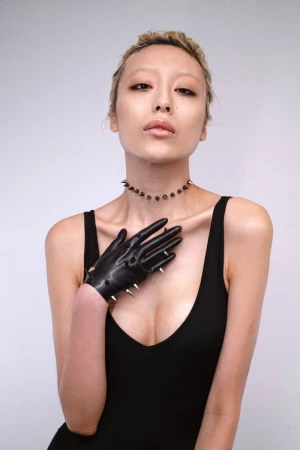 Woman in black dress and spiked glove posing in a studio with a light grey background, wearing a spiked necklace, eye-level, medium shot.