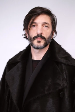 Portrait of an adult male with a beard, wearing a black fur coat against a white backdrop in a studio environment.