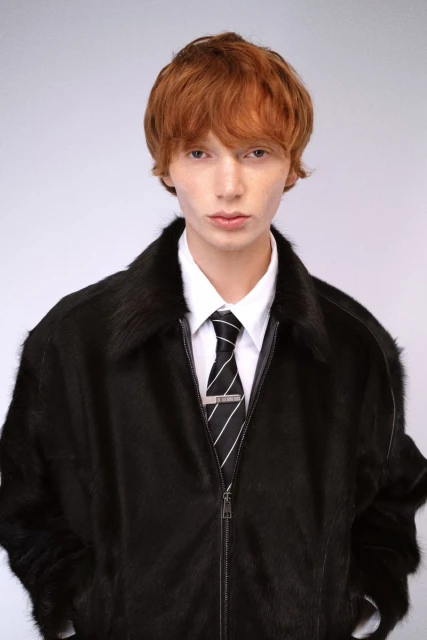 Close-up of a red-haired adult male in a black fur jacket, white shirt, and striped tie, posing in a photo studio with a light gray backdrop.