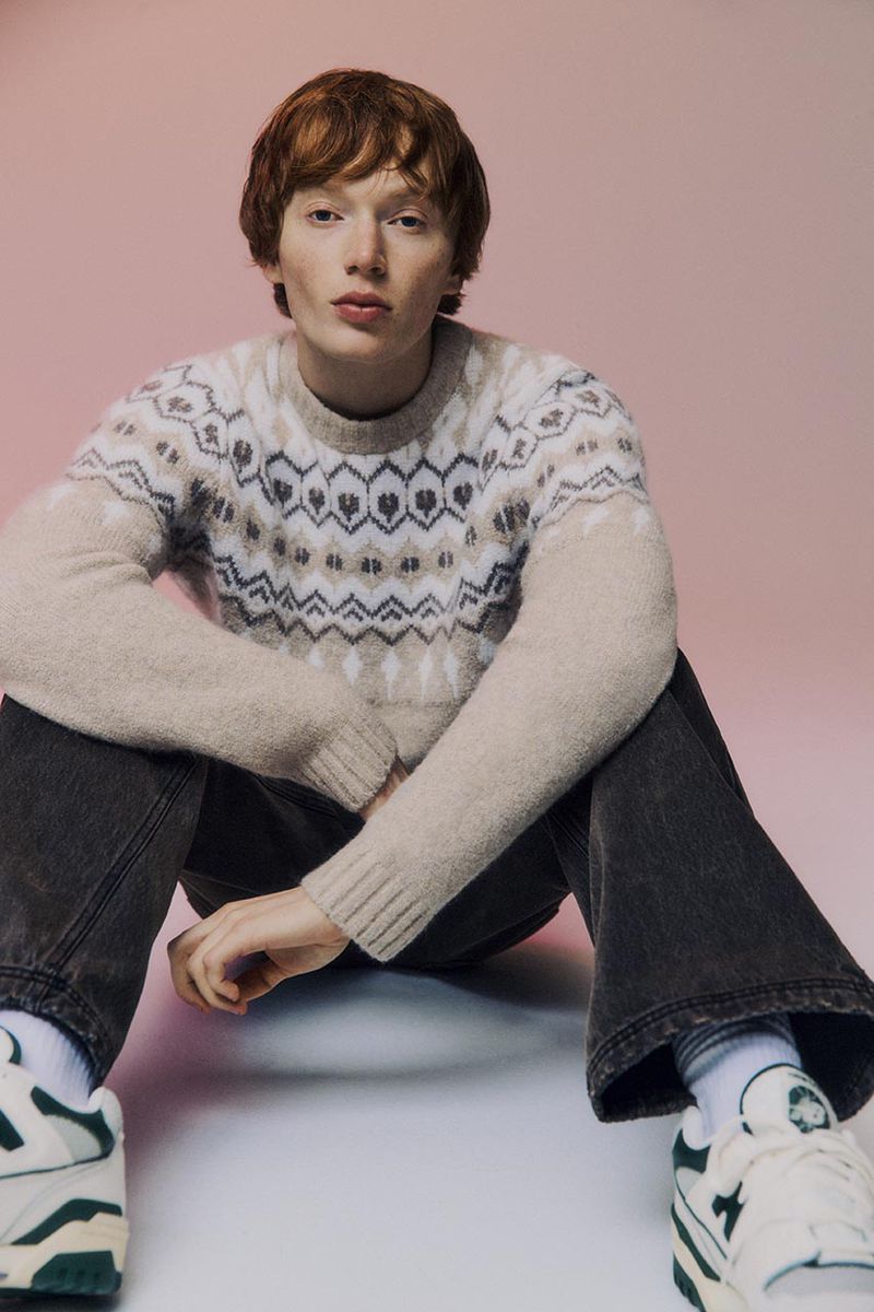 A young adult man with red hair sits in a studio setting wearing a beige patterned sweater, dark jeans, and athletic sneakers.