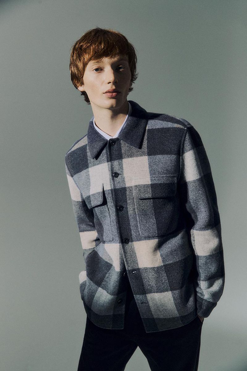 A young adult male model poses indoors wearing an open, grey and beige plaid button-down shirt over a white t-shirt and black pants.
