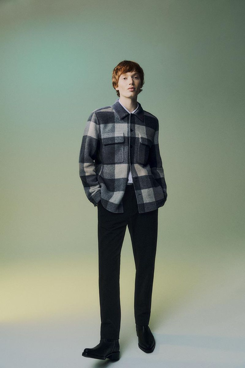 Full body shot of a young adult male with auburn hair wearing a gray and black plaid flannel shirt, black pants and boots, in a studio setting.