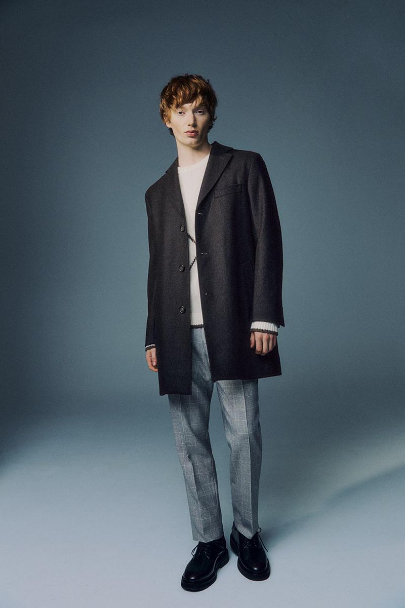 Male fashion model posing in studio wearing dark overcoat, sweater, plaid pants, and black shoes against a gradient blue background.