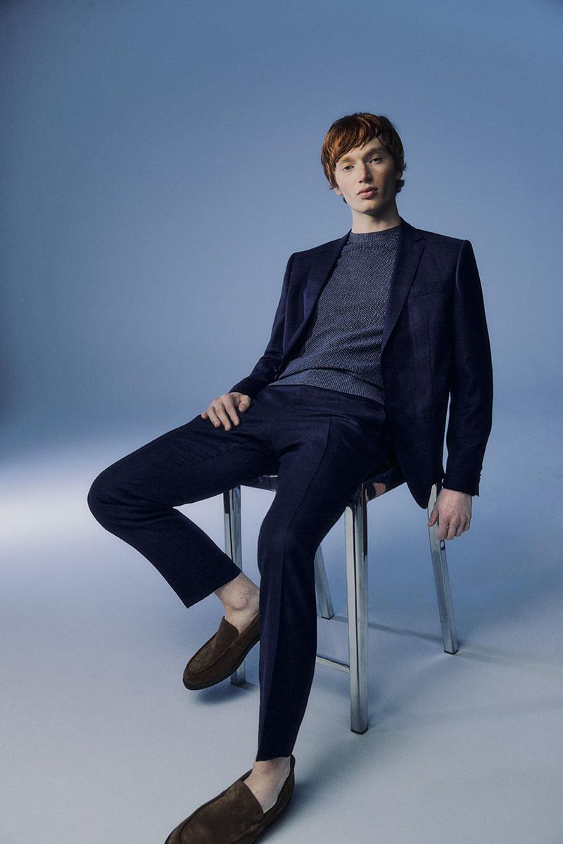 A stylish adult male with red hair in a blue suit and brown loafers, sitting on a stool against a blue backdrop in a studio setting.