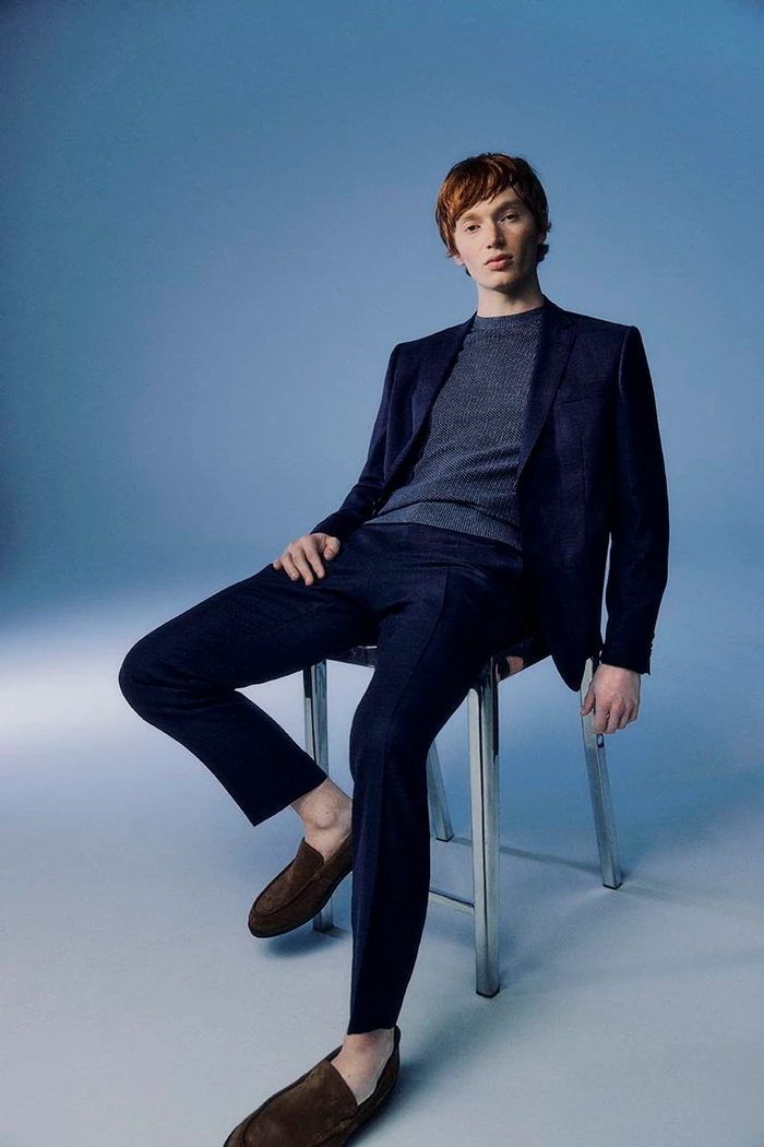 A red-haired male model seated on a chrome chair in a navy suit against a blue studio background for Nordstrom Holiday 2024.