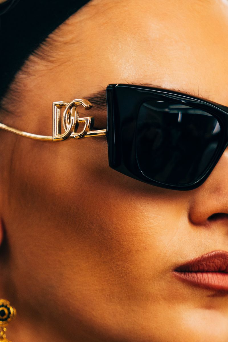 Close-up of a fair-skinned person wearing square black sunglasses with a gold DG logo on the frames, showcasing high-fashion eyewear.
