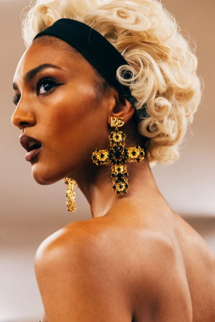 Close-up portrait of a woman with blonde curly hair and a black headband wearing large, gold cross-shaped earrings.