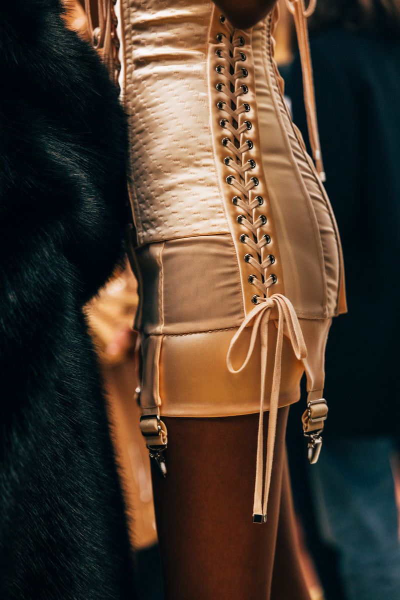 Close-up of a person wearing a tan corset with lace-up detailing and attached suspender straps, alongside a black fur garment.