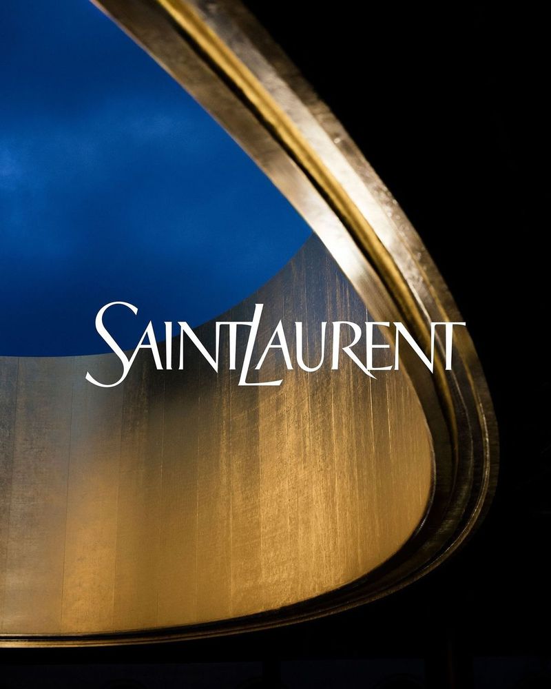 Promotional image featuring a golden curved structure with the 'Saint Laurent' logo against a dark black and blue background.