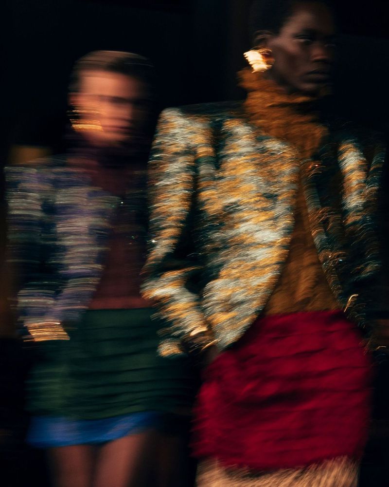Two fashion models in motion, showcasing a green mini skirt and a patterned jacket with gold accents, captured in a dynamic blur against a dark backdrop.