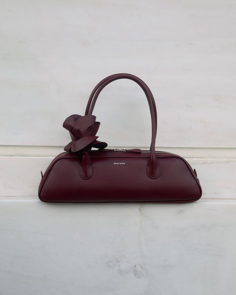 Burgundy leather handbag with a matching leather flower accessory, displayed against a white textured surface in a studio setting.