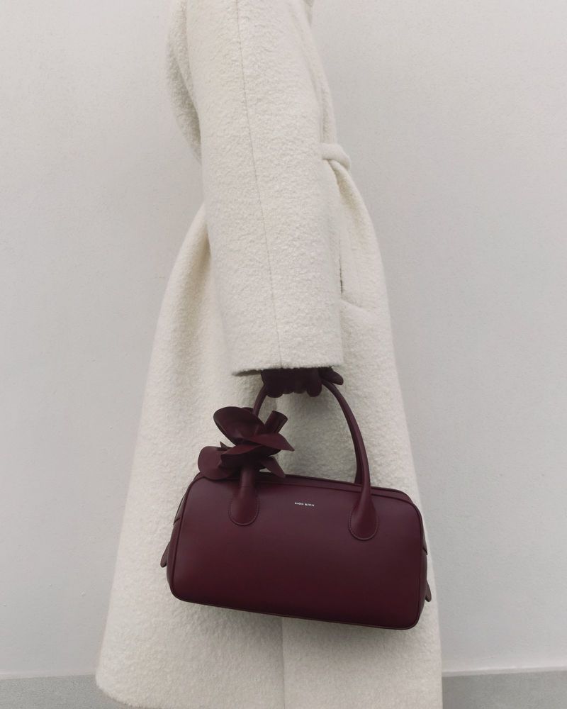Burgundy handbag with floral detail carried by a person wearing a white wool coat against a neutral background.