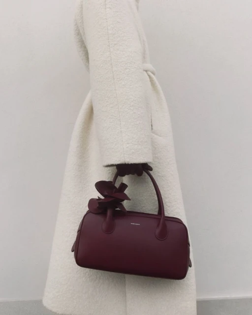 Burgundy handbag with floral detail carried by a person wearing a white wool coat against a neutral background.