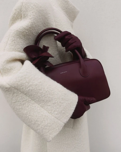 Burgundy satchel handbag with a bow, held by a person wearing matching gloves and a textured off-white coat, on a neutral background.