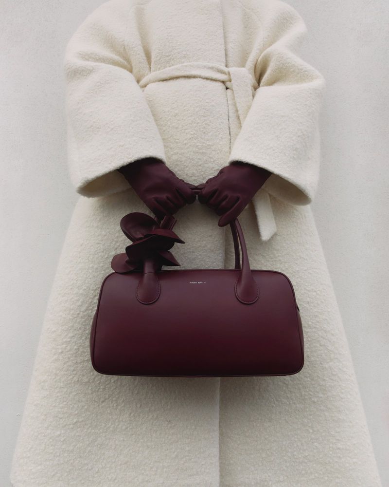 Burgundy handbag with flower detail held by adult in cream-colored belted coat and maroon gloves against an off-white background.