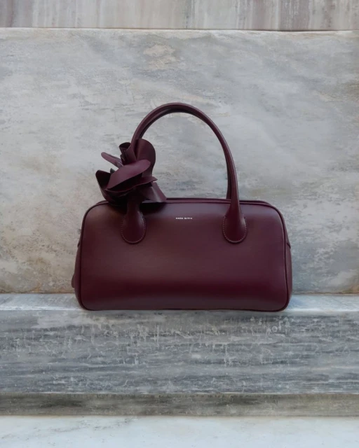 Burgundy leather handbag with top handles and a decorative flower accent, presented against a textured gray background. Fashion accessory.