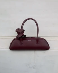 Burgundy leather handbag with structured handles and a matching flower appliqué resting against a white marble surface.