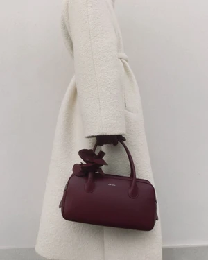 Model wearing white textured coat holding a structured burgundy handbag by Magda Butrym against a plain studio background.