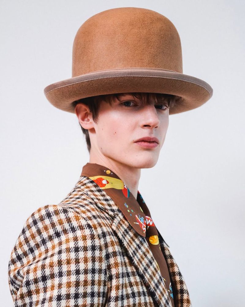 Portrait of a young adult male model wearing a tan bowler hat, brown patterned blazer, and brown patterned scarf against a white background.