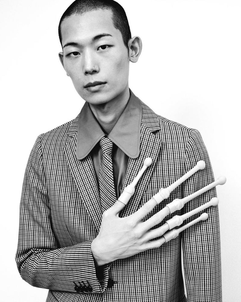 Monochrome studio shot of an Asian male model in patterned suit with claw-like finger extensions against a white background, portrait.