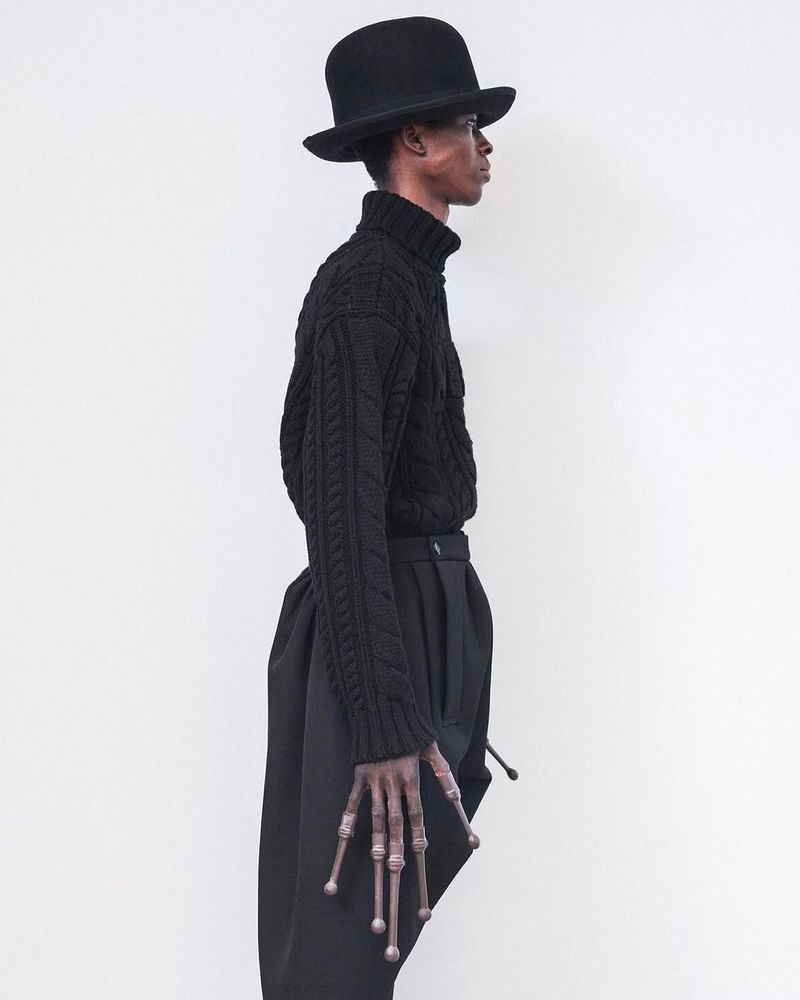 Dark-skinned adult posing indoors, wearing a black bowler hat, cable knit sweater, and wide-legged pants, with decorative hand extensions.