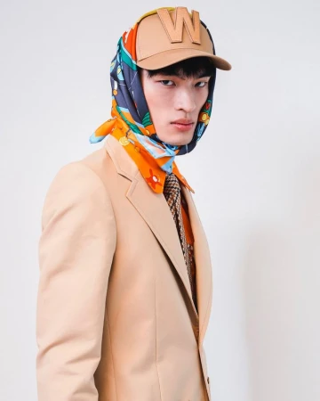 Fashion portrait of a young Asian adult male wearing a beige suit, cap, and patterned headscarf in a bright studio setting.