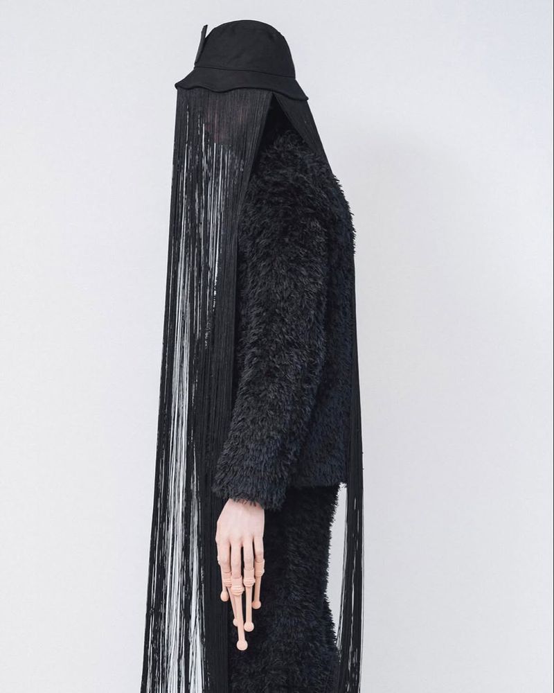Black fashion outfit featuring a fringed hat, textured jacket, and hand with ring details, set against a plain white background.