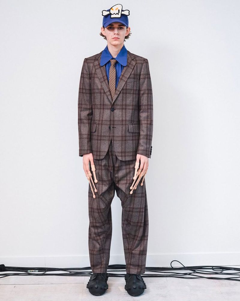 Full-length studio shot of a young adult man wearing a plaid suit, blue collared shirt, tie, cap with drumsticks attached to his pants.