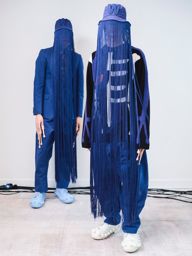 Two people in matching blue outfits with fringe masks and bucket hats stand indoors on a pale surface with cables.