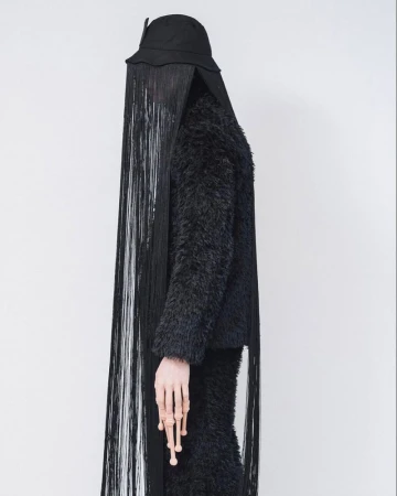 Model wearing a black furry textured jacket, black bucket hat, and long fringe veil covering the face against a white background
