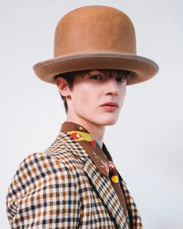 Model wearing a brown felt bowler hat and patterned jacket against a white background