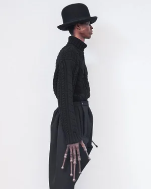 Profile view of a black man wearing a black bowler hat, turtleneck sweater, and prosthetic fingers against a white background