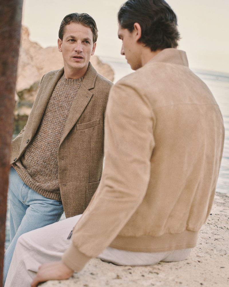 Two men in stylish jackets stand near the water; one looks to the left, the other faces away in a close outdoor shot.