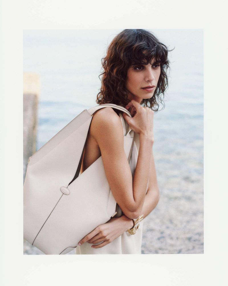 A fashionable woman with a shoulder bag stands outdoors, showcasing the handbag. She wears a light dress and a gold bracelet.