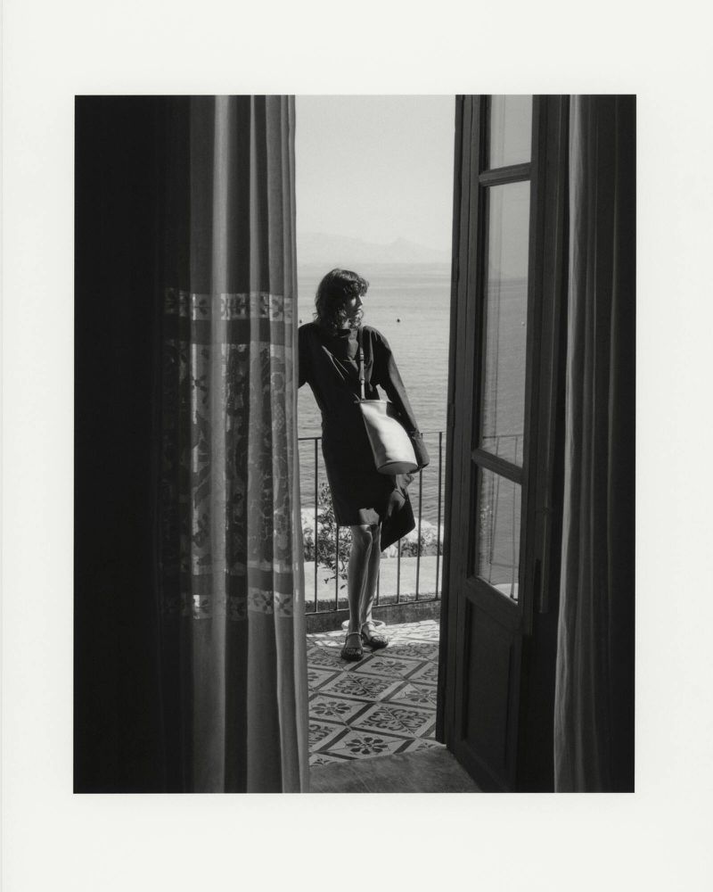 Black and white shot of a woman standing on a balcony overlooking the sea, visible through a doorway framed by draped curtains.